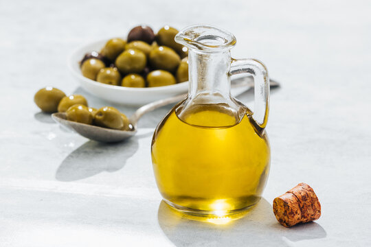 Glass jug with oil and small bowl and spoon with olives placed on white table