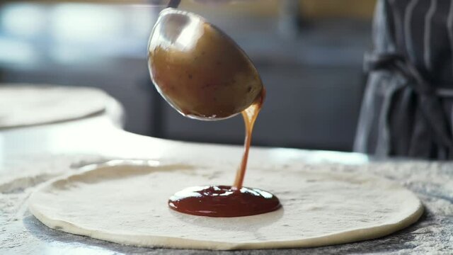 Close Up of Chef is spreading Tomato Sauce on Pizza Dough with a Metal Ladle in Traditional Italian Pizza's Restaurant. Cooking traditional Sause for Italian Pizza. Italian Food. Pizza's Making.