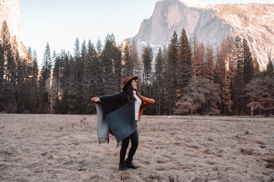 Happy relaxed young female traveler in stylish outfit sitting on stone border against picturesque mountain scenery with rocky cliffs and coniferous forest in Yosemite National Park in USA