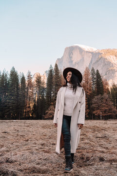 Happy relaxed young female traveler in stylish outfit sitting on stone border against picturesque mountain scenery with rocky cliffs and coniferous forest in Yosemite National Park in USA