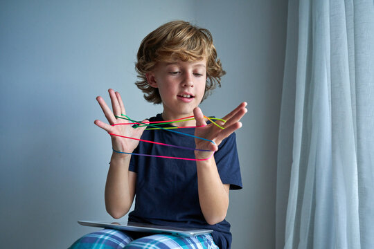 Concentrated preteen boy in pajama sitting on wooden stool with tablet on knees and creating string figure while playing creative cats cradle game at home