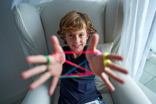 Closeup of hands of boy playing cats cradle game and demonstrating basic figure made with colorful string while spending free time at home looking at camera