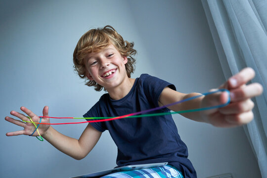 Excited schoolboy in pajamas with tablet on knees creating difficult figure with string while playing cats cradle game at home sitting on stool