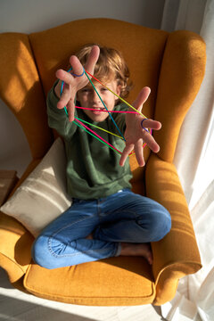From above cheerful preteen boy in casual clothes sitting in cozy armchair and playing interesting cats cradle game while resting at home looking at camera