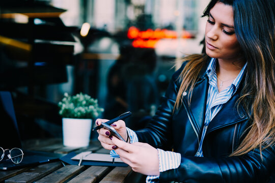 Cropped image of attractive female blogger with long hair publishing new post in social networks on modern mobile phone using free internet connection sitting outdoors in urban setting.Copy space area