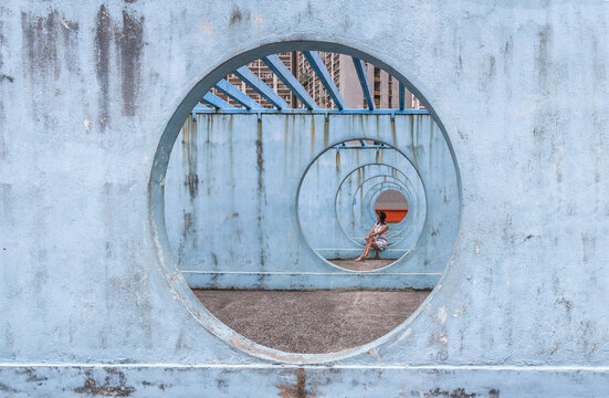 Female in summer dress sitting on concrete base of unusual city installation in shape of passage with round holes and looking away