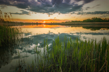 Scenic view at beautiful spring sunset with reflection on a shiny lake with green reeds, bushes, grass, golden sun rays, calm water ,deep blue cloudy sky , glow on background, spring evening landscape