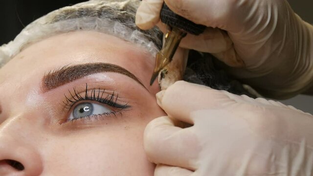 A special needle tattoo machine makes permanent makeup correction of a young woman's eyebrows. A pigment of dark paint is injected under the skin. Microblading, powder spraying.