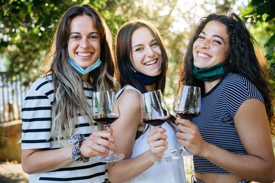 Group of young millennials women having fun in a park drinking red wine at a party with the face mask lowered under the chin avoid Coronavirus, Covid-19 - Friends meet after the lockdown at appetizer