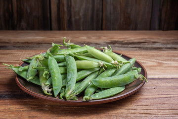 green pea pods in a clay plate on a wooden table close-up, harvest from the garden