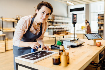 Brunette tired woman owner of zero waste shop.Female worker in grey apron preparing , writing open...