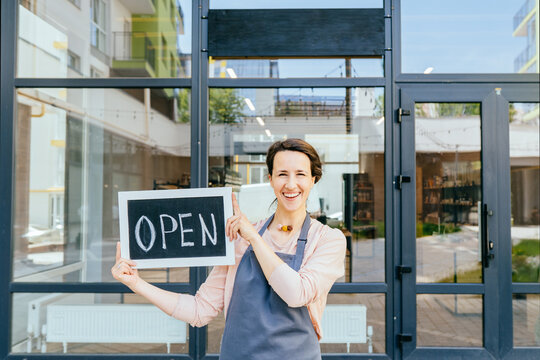 Portrait of positive excited young charming woman holding open sign over shop facade. Female owner local business enjoying opening after coronavirus is outbreak in city.