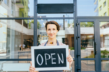 Close up portrait of french saleswoman or owner business woman showing open sign standing against the door frame of an organic or zero waste store. Opening after coronavirus outbreak in city.