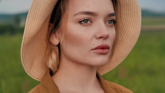Portrait of Beautiful Country woman standing at Empty green Field, feeling of absolute Freedom. Looking sad and Confident. Having trendy Straw hat and wearing light clothes. Summer evening.