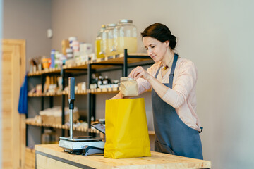 Young smiling positive female volunteer collecting food donations into paper bags indoors. Positive woman owner in apron standing behind counter in package free grocery store collects paper bag.