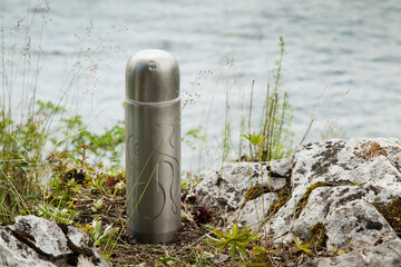 A tourist steel thermos with an engraving stands on the Bank of the river.