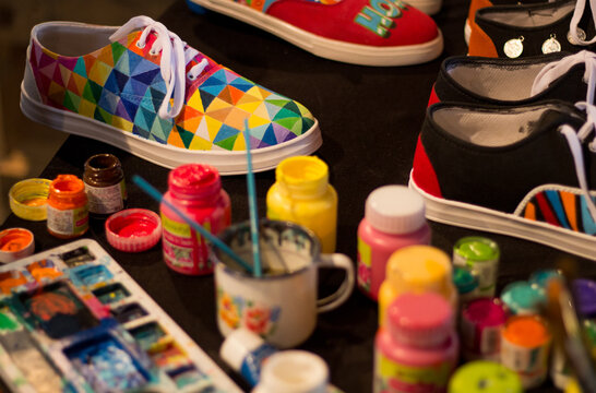 A fresh colorfully hand-painted shoe kept on a desk along with other colorful shoes, paints, and brushes.