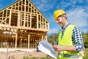 Engineer with hardhat and blueprints on building site of wood frame house under construction