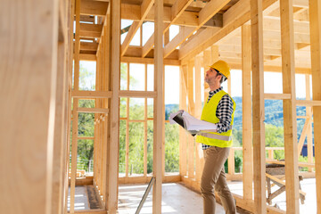 Engineer with hardhat and blueprints on building site of wood frame house under construction