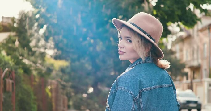 Beautiful blonde Woman with trendy Hat having a walk at Sunset in the Street. Looking back , at the Camera. Having nice Appearance, casually dressed. Green trees and pleasant weather outdoor.