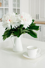 White peonies in a vase and a cup of milk on a white background in the interior. Breakfast. Composition.