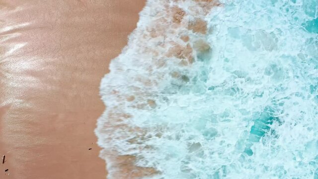 Aerial top view of ocean blue waves break on a beach. Sea waves and beautiful sand beach aerial view drone shot. Bird's eye view of ocean waves crashing against an empty sand beach from above.