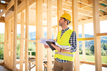 Construction engineer or architect with blueprints visiting building site of wood frame house