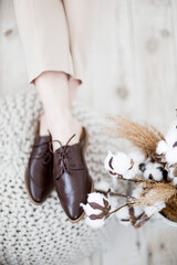 fashionable women's shoes, feet in the interior, classic women’s flat shoes, brown leather shoes, mules and loafers, knitted blankets and blankets on the background, wooden background, cotton flowers,