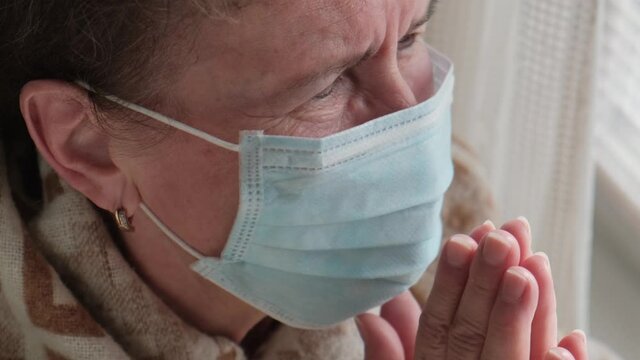 An elderly woman in a protective mack is praying to god by the window during a coronavirus pandemic