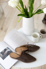 leather women’s flat shoes with laces, brown mules and trendy loafers, in the interior, lie on the table, flowers, magazine, morning, atmosphere, tea cup, product display lifestyle