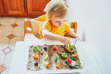 Adorable little girl playing with toy fruits and vegetables and kinetic sand at home