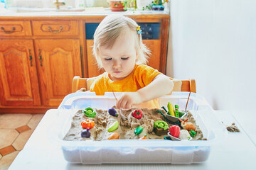 Adorable little girl playing with toy fruits and vegetables and kinetic sand at home