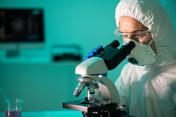 Concentrated virologist in safety goggles and respiratory mask using microscope in dark laboratory while researching virus cells
