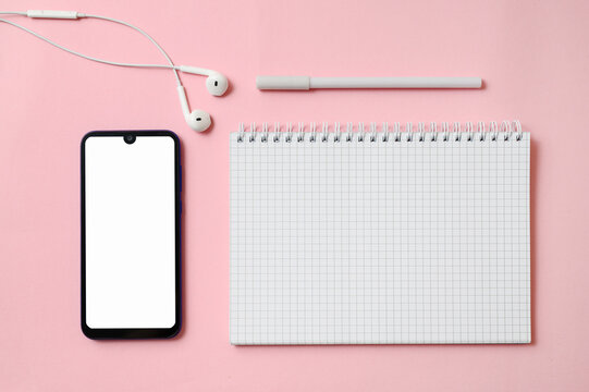 notebook pen earphones and smartphone with white display lie on a pink background