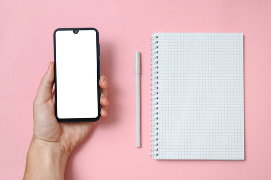 Male hand holds a smartphone with a white display. Next to the hand lies a notebook with a blank page and pen for writing. Pink background.