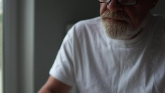 A sad gray-bearded man with glasses shakes his head, counts cash using a calculator in a smartphone. Financial crisis, falling income