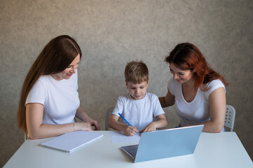 Happy same-sex lesbian family with a child stays at home. Two mothers help the boy do school homework. A couple of women help their son with distance learning. Quarantine.