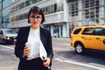 Half-length portrait of businesswoman in formal outfit waiting for taxi holding coffee to go cup...