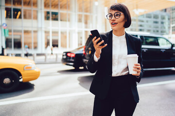 Thoughtful female economist in trendy eyeglasses holding mock up coffee to go cup and looking away standing on street.Trader in elegant formal wear strolling in downtown with telephone in hand