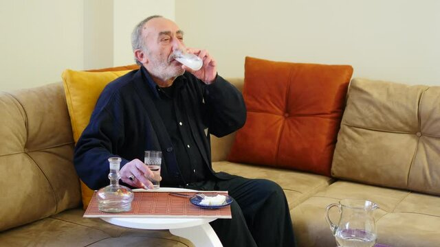 Old man drinks Raki and eats White Cheese. Senior sits on the sofa in living room. If you like liqueur and anise, you will definitely love the taste of Raki alcoholic drink, made of grapes and raisins