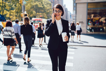 Half-length portrait of businesswoman in formalwear passing crosswalk talking on phone satisfied with news, prosperous female trader crossing avenue in sunny day making phone call to share good news.