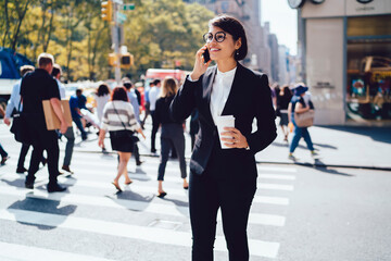 Positive corporate manager talking on phone during coffee break outdoors holding takeaway cup, businesswoman in formal wear and eyewear enjoying rest having mobile conversation with colleague