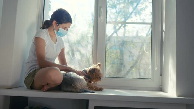 coronavirus pandemic concept. little teenage girl sitting in a medical mask at the window with a shaggy dog. self-isolation concept virus covid 19 infection doomsday. Sick child kid indoors. girl