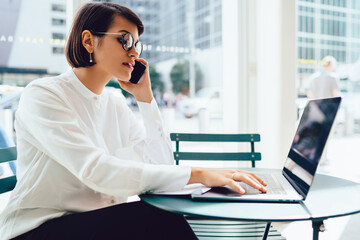 Serious female lawyer in eyeglasses booking tickets during mobile conversation sitting at laptop device with wireless internet connection.Businesswoman talking on cellular while doing remote job