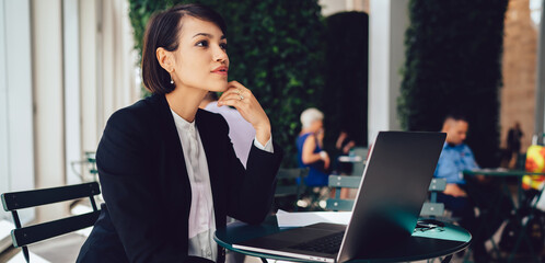 Contemplative businesswoman dressed in elegant formal black suit thoughtful looking away while...