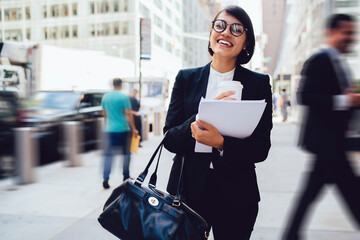 Half length portrait of excited businesswoman laughing on modern streets in metropolis during work break for coffee time, happy cheerful female in elegant suit holding takeaway cup with beverage