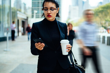 Serious female manager in elegant suit making payment on online service using 4g internet on cellphone during way to office, trendy woman employee checking notification on modern smartphone device