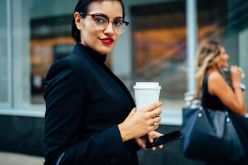 Portrait of confident female manager in stylish eyewear using smartphone for banking standing outdoors, serious businesswoman with red lips holding coffee to go cup looking at camera