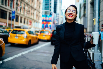 Naklejka premium Cheerful financial expert with coffee to go laughing on modern street while waiting for taxi cab near road with traffic on Manhattan, happy excited businesswoman in optical spectacles feeling good