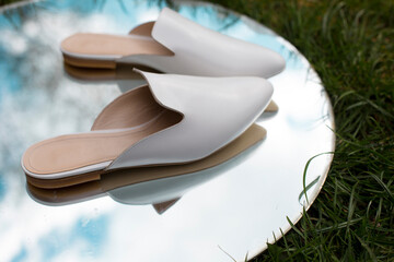 white casual leather slippers on the street, women’s flat shoes comfortable and useful lie on a fashionable round mirror with reflection of shoes, sky and grass